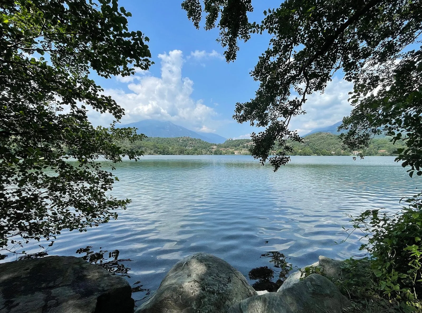 Il Lago Sirio vicino a Ivrea visto dalla riva, tra rami di alberi, con colline verdi e montagne sullo sfondo