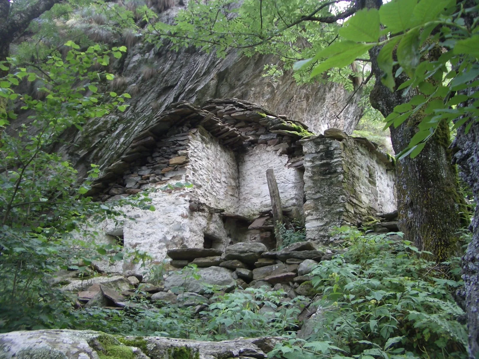 Antica baita in pietra con il tetto in lose addossata a una parete di roccia nel bosco della Valchiusella